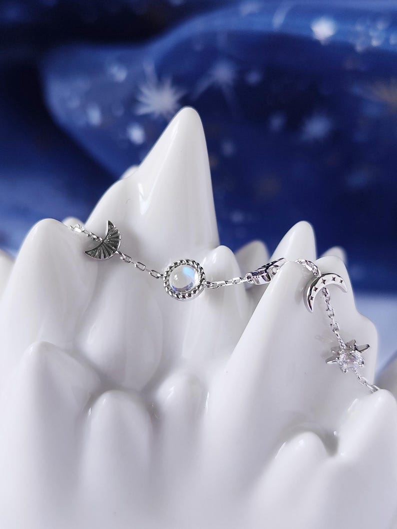 May include: A silver chain bracelet with a moon and star charm, a round iridescent gemstone, and a crescent moon charm.
