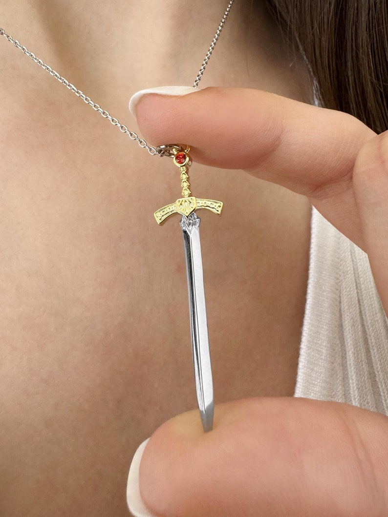 May include: A silver chain necklace with a gold and silver sword pendant. The sword has a red gemstone at the top.