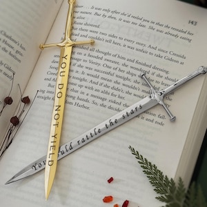 Puede incluir: Dos marcadores de libros con forma de espada, uno de oro y otro de plata. El marcador de oro tiene el texto "You Do Not Yield" grabado. El marcador de plata tiene el texto "You Should Rattle The Stars" grabado. Los marcadores est&aacute;n sobre un libro con un helecho y una pi&ntilde;a.
