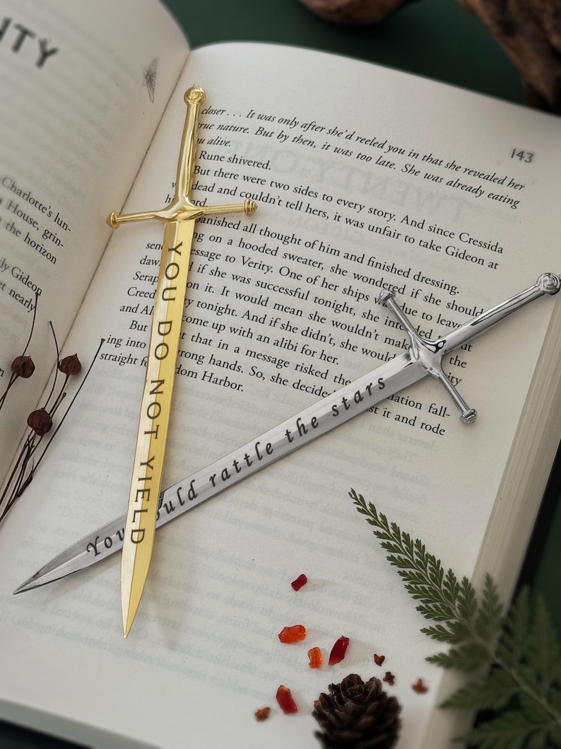 Puede incluir: Dos marcadores de libros con forma de espada, uno de oro y otro de plata. El marcador de oro tiene el texto "You Do Not Yield" grabado. El marcador de plata tiene el texto "You Should Rattle The Stars" grabado. Los marcadores est&aacute;n sobre un libro con un helecho y una pi&ntilde;a.