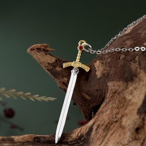 May include: A silver and gold sword pendant necklace with a red gemstone on the hilt. The pendant is hanging from a silver chain.
