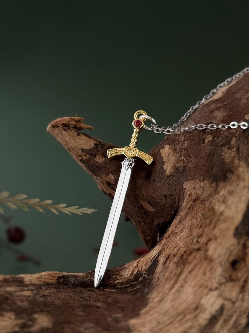 May include: A silver and gold sword pendant necklace with a red gemstone on the hilt. The pendant is hanging from a silver chain.