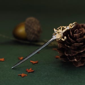 May include: A miniature sword pendant with a gold-colored hilt and a silver blade. The hilt features intricate leaf and floral designs, with a small gemstone. The pendant rests on a dark green surface with scattered orange leaves.