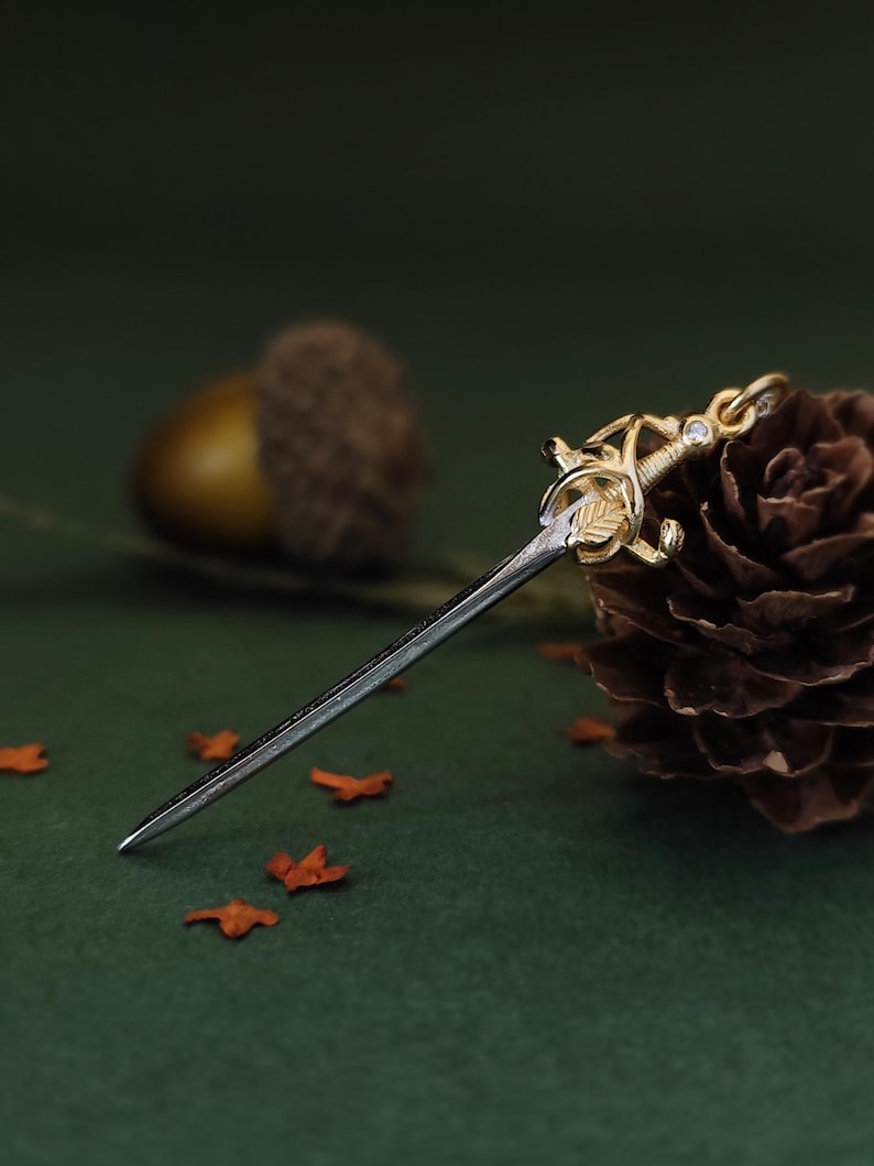 May include: A miniature sword pendant with a gold-colored hilt and a silver blade. The hilt features intricate leaf and floral designs, with a small gemstone. The pendant rests on a dark green surface with scattered orange leaves.