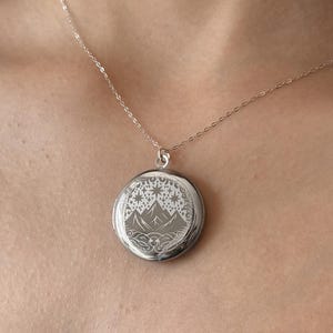 May include: A silver locket necklace with a mountain and stars design. The locket is round and has a delicate chain.