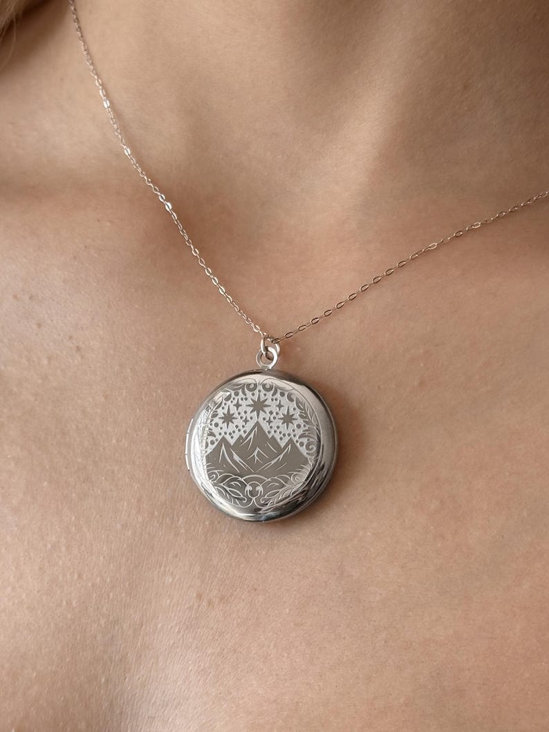 May include: A silver locket necklace with a mountain and stars design. The locket is round and has a delicate chain.