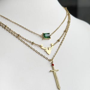 May include: A gold layered necklace with three pendants: a green square gem, a deer silhouette, and a sword with a red gemstone.