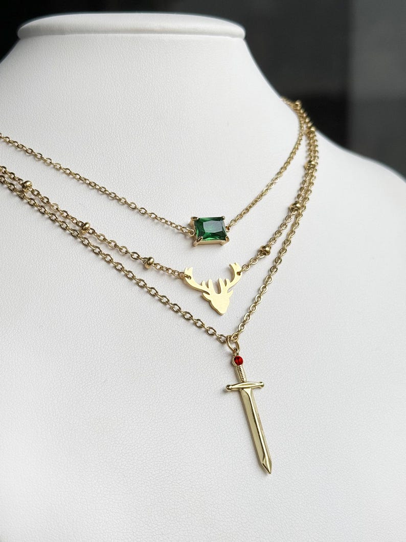 May include: A gold layered necklace with three pendants: a green square gem, a deer silhouette, and a sword with a red gemstone.