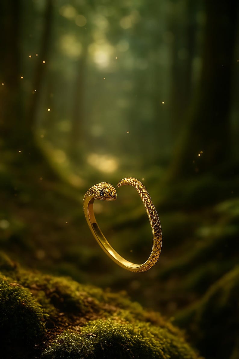 May include: A gold-colored snake ring with a textured pattern. The snake's head features a small, dark gemstone eye. The ring is set against a blurred green background, suggesting a natural setting.