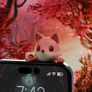 Puede incluir: Un charm de teléfono de zorro rosa de dibujos animados que sostiene una manzana roja. El charm está sentado encima de un teléfono inteligente negro con una esfera de reloj que marca las 7:43. El fondo es una imagen borrosa de un bosque rojo.