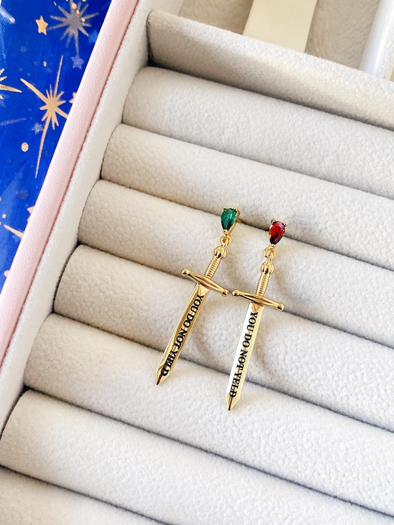 May include: Two gold sword earrings with a teardrop-shaped green and red stone at the top. The swords have the words "You Do Not Yield" engraved on them.