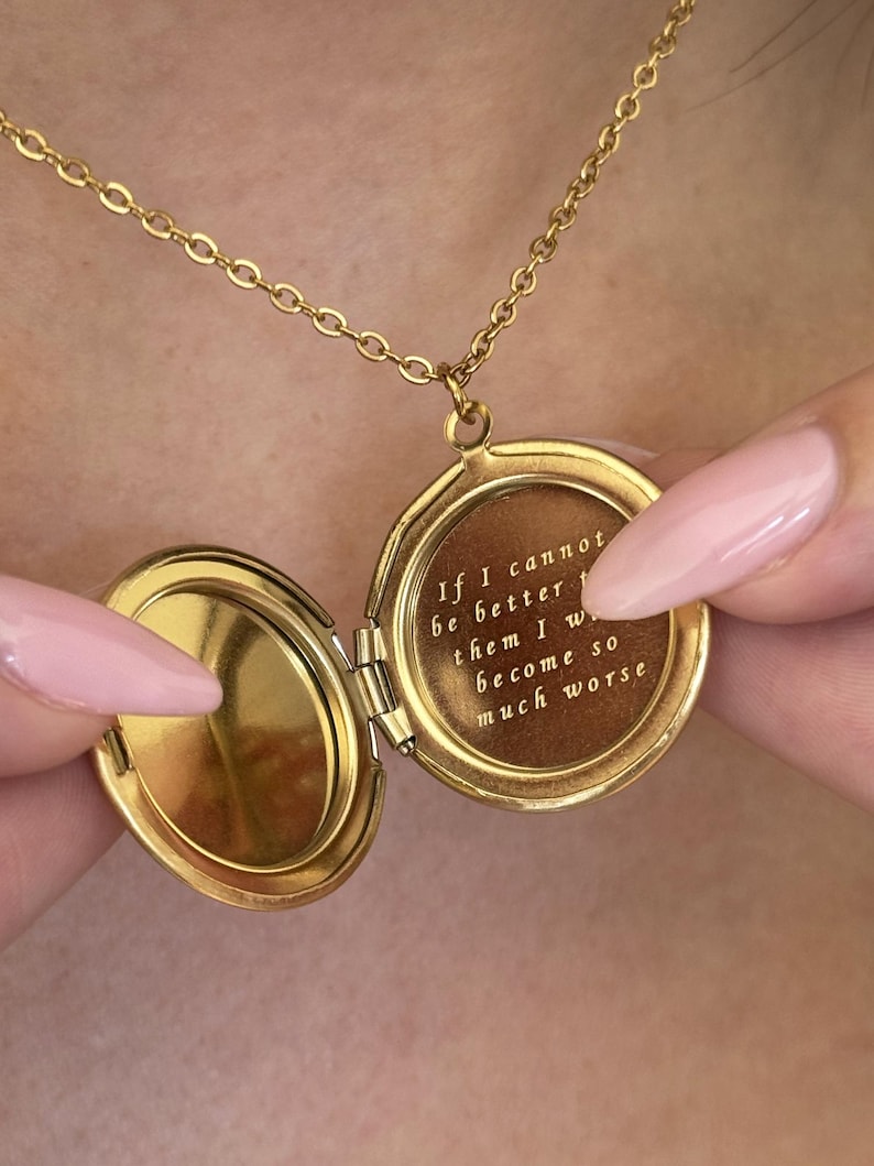 May include: A gold-colored locket necklace with an open, round pendant. The pendant reveals engraved text: "If I cannot be better than them I will become so much worse."