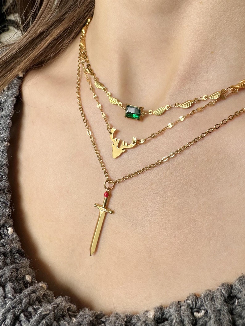 May include: Three gold necklaces with pendants. The top necklace has a green gemstone pendant. The middle necklace has a deer head pendant. The bottom necklace has a sword pendant with a red jewel.
