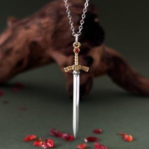 May include: A silver chain necklace with a gold and silver sword pendant. The sword has a red gemstone at the top.
