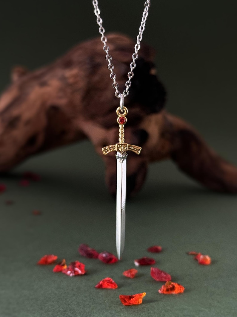 May include: A silver chain necklace with a gold and silver sword pendant. The sword has a red gemstone at the top.