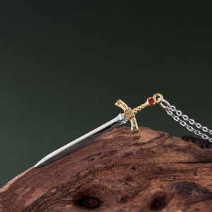 May include: A silver and gold sword pendant necklace with a red gemstone on the hilt. The necklace is laying on a piece of weathered wood.