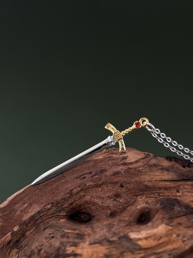 May include: A silver and gold sword pendant necklace with a red gemstone on the hilt. The necklace is laying on a piece of weathered wood.
