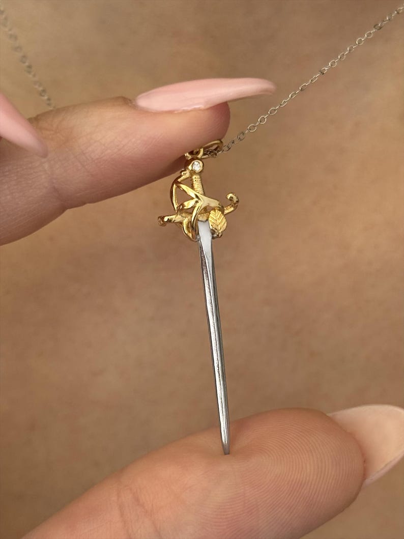 May include: A gold-colored pendant necklace featuring a miniature sword design. The sword has a silver blade and is adorned with gold-colored floral and leaf details. A small diamond is set at the top of the sword's hilt.