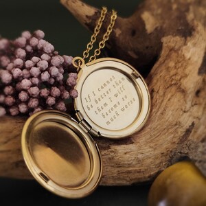 May include: A gold-colored locket necklace, open to reveal an engraved quote. The locket hangs on a delicate chain. The quote reads: "If I cannot be better than them I will become so much worse."