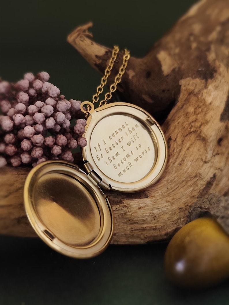 May include: A gold-colored locket necklace, open to reveal an engraved quote. The locket hangs on a delicate chain. The quote reads: "If I cannot be better than them I will become so much worse."