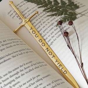 Puede incluir: Un marcap&aacute;ginas de oro con forma de espada con las palabras "You Do Not Yield" grabadas en &eacute;l. El marcap&aacute;ginas est&aacute; sobre un libro abierto con una hoja de helecho y flores secas.