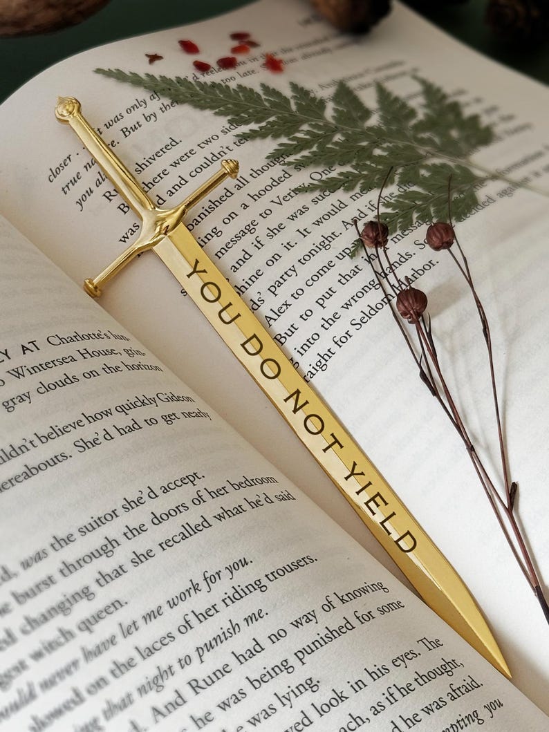 Puede incluir: Un marcap&aacute;ginas de oro con forma de espada con las palabras "You Do Not Yield" grabadas en &eacute;l. El marcap&aacute;ginas est&aacute; sobre un libro abierto con una hoja de helecho y flores secas.
