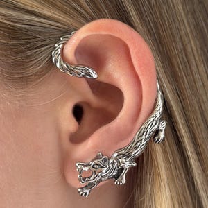 May include: Silver-toned ear cuff featuring a detailed wolf design. The cuff wraps around the ear, with the wolf's head positioned near the earlobe and its body extending along the outer ear. The jewelry is intricately carved.