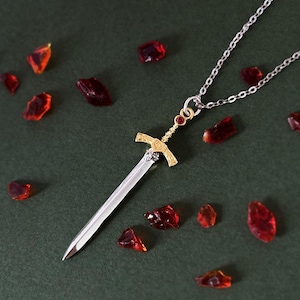 May include: A silver chain necklace with a gold sword pendant. The sword has a red gemstone at the top and is surrounded by red glass shards.