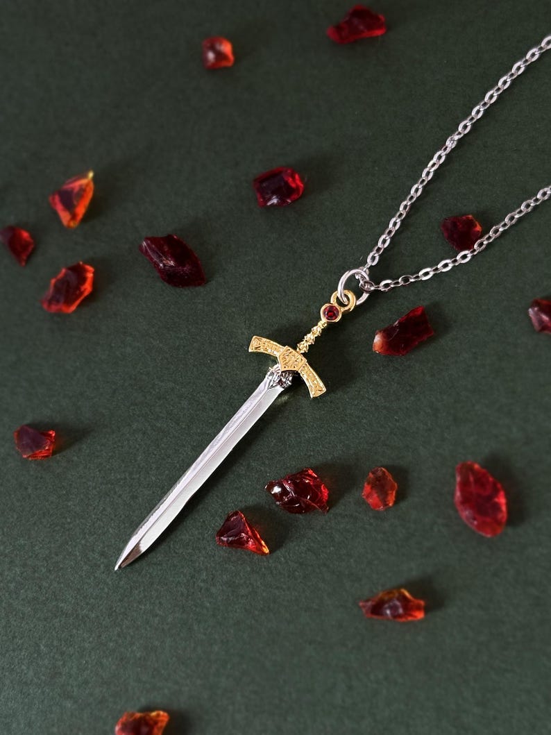 May include: A silver chain necklace with a gold sword pendant. The sword has a red gemstone at the top and is surrounded by red glass shards.