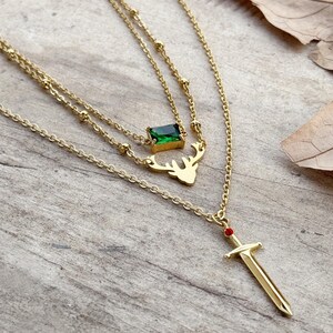 May include: Gold layered necklace with a green gemstone, a deer pendant, and a sword pendant with a red gemstone.