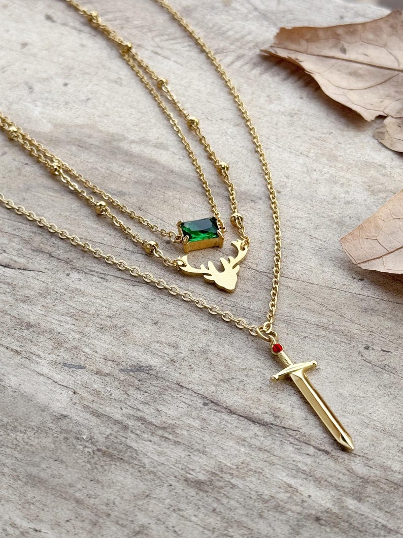 May include: Gold layered necklace with a green gemstone, a deer pendant, and a sword pendant with a red gemstone.