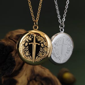 May include: Two round pendant necklaces. One is gold-toned, the other silver-toned. Both feature a sword design within a circular frame. The gold necklace has a black background, while the silver one has a silver background. Both hang from delicate chains.