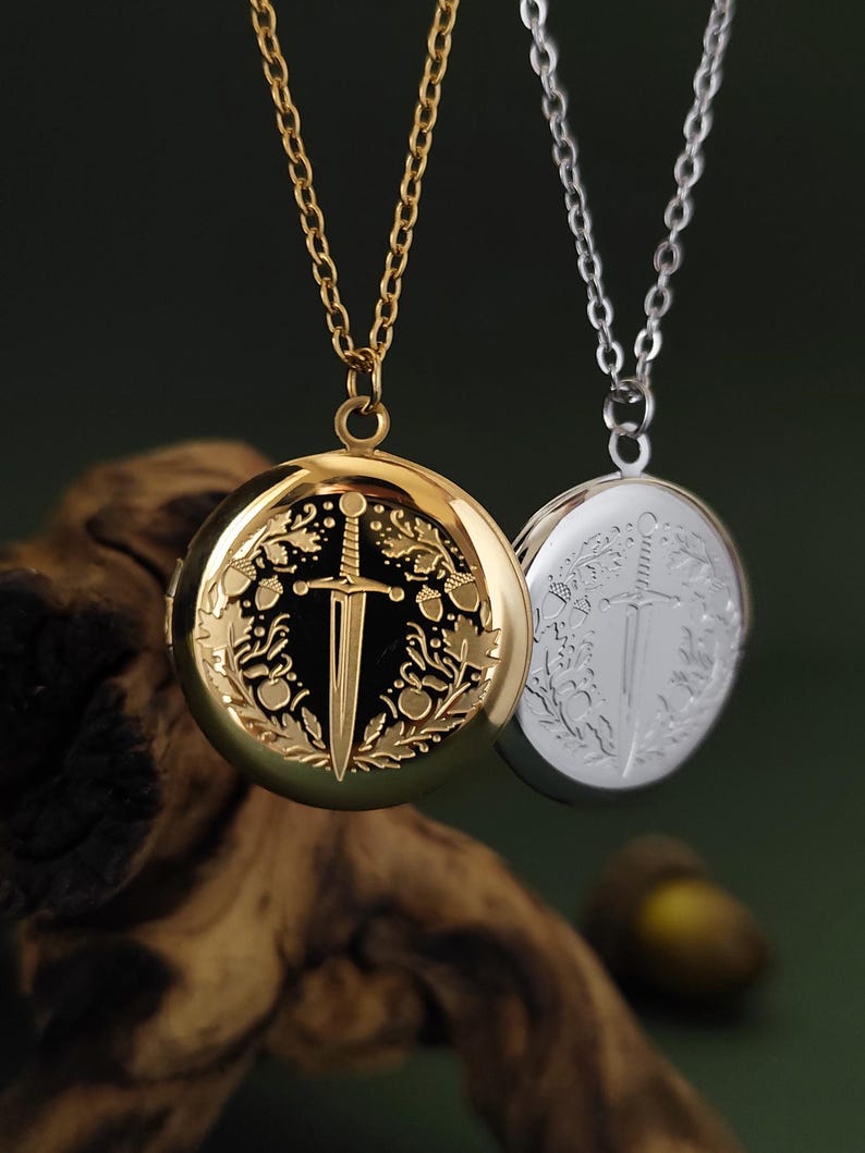 May include: Two round pendant necklaces. One is gold-toned, the other silver-toned. Both feature a sword design within a circular frame. The gold necklace has a black background, while the silver one has a silver background. Both hang from delicate chains.
