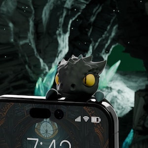 Op de afbeelding: Een zwarte telefoon met een donker achtergrondbeeld op het scherm. De tijd is 7:43. Een klein zwart drakenbeeldje met gele ogen zit bovenop de telefoon.