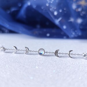 May include: A silver chain bracelet with a moonstone charm and crescent moon charms. The bracelet is on a white surface.