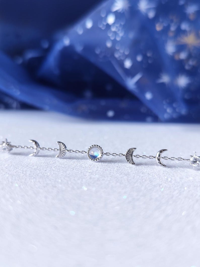 May include: A silver chain bracelet with a moonstone charm and crescent moon charms. The bracelet is on a white surface.