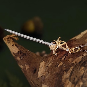 May include: A gold and silver pendant necklace featuring a miniature sword design. The sword has a detailed hilt with a leaf motif and a silver blade. The necklace is displayed on a piece of wood against a blurred green background.