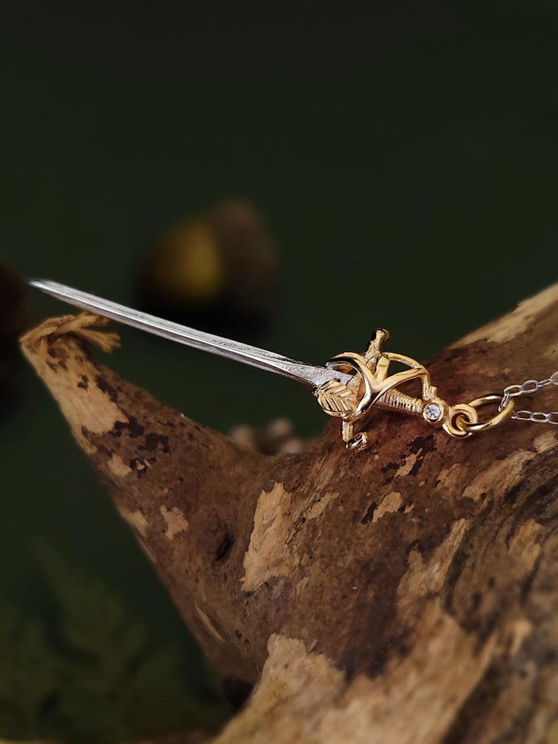 May include: A gold and silver pendant necklace featuring a miniature sword design. The sword has a detailed hilt with a leaf motif and a silver blade. The necklace is displayed on a piece of wood against a blurred green background.