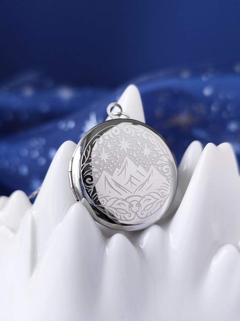 May include: A silver locket necklace with an engraved design of a mountain range and stars. The locket is round and has a hinged lid.