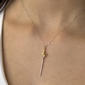 May include: A gold-colored sword pendant necklace with a silver chain. The pendant features a detailed hilt and a slender blade. The necklace is worn against a person's skin.