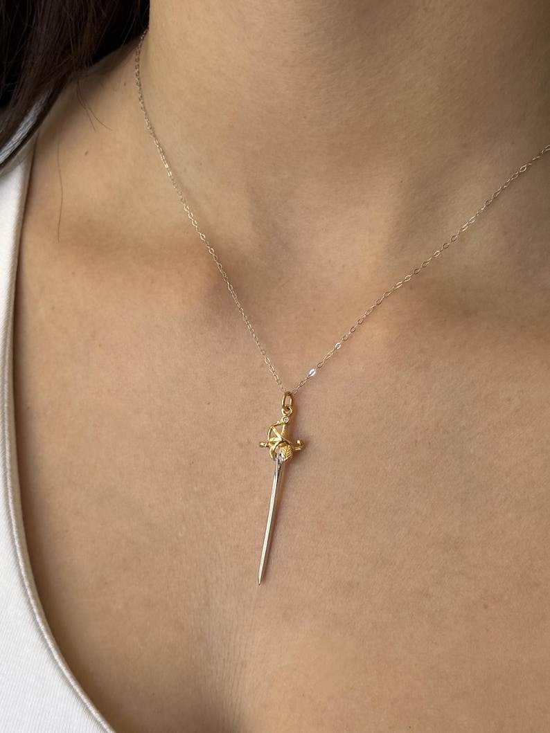 May include: A gold-colored sword pendant necklace with a silver chain. The pendant features a detailed hilt and a slender blade. The necklace is worn against a person's skin.