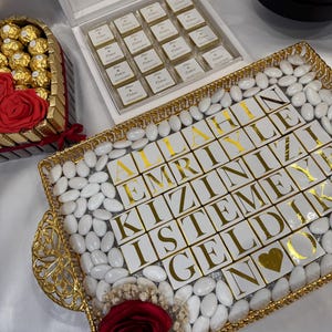 May include: A gold tray filled with white almond-shaped candies and white tiles with gold lettering spelling out "ALLAHIN EMRIYLE KIZINIZI ISTEMEY GELDIK NOU" with a gold heart in the center. A red rose is in the corner of the tray.