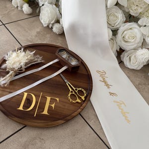 May include: A wooden wedding ring tray with gold initials "D/F" and a pair of gold scissors. A small wooden box with rings is on the tray. A white ribbon and white flowers are in the background.