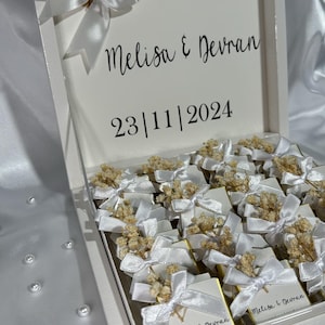 May include: White box with a white ribbon and dried flowers on top. The box is filled with small white boxes with white ribbons and dried flowers. The box is labeled "Melisa & Devran 23|11|2024".