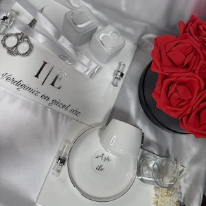 May include: A white and silver gift set with a coffee cup and saucer, two ring boxes, and a decorative tray. The tray has text in a script font. A bouquet of red roses is in the background. The set is on a white satin surface.