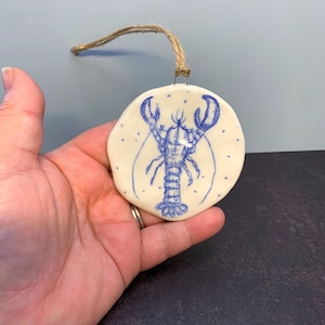 May include: A round, off-white ceramic ornament featuring a detailed blue line drawing of a lobster in the centre. The ornament has an irregular, hand-formed edge and is suspended by a thin, natural-coloured jute twine loop attached at the top.