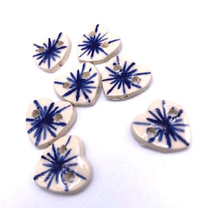 May include: A selection of heart-shaped buttons. Each button is off-white with a blue starburst design and two holes for sewing. The buttons are made of ceramic.