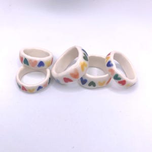 May include: Several white ceramic rings decorated with colourful heart motifs. The hearts are in red, yellow, green, and blue. These rings offer a playful and bright aesthetic.