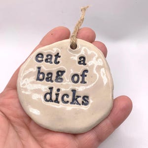 May include: A cream ceramic ornament with the words "eat a bag of dicks" in dark blue. The ornament has an irregular shape and a jute twine loop for hanging. A humorous gift.