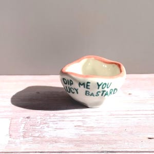 May include: A small, white ceramic bowl with a pink rim and the text "DIP ME YOU LUCY BASTARD" in green lettering. The bowl has an irregular shape and is sitting on a light-colored wooden surface.